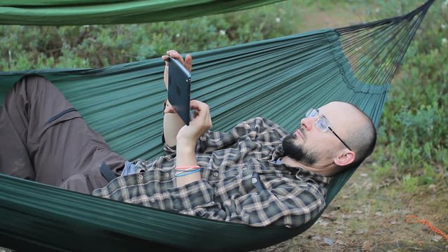 A man in a hammock touch tablet. Man in the woods with a beard and glasses