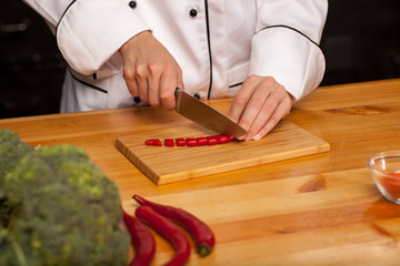 Female chef cook cutting pepper on wooden cutting board, top view. Broccoli, salad leaves, tomatoes and pepper on the table.
