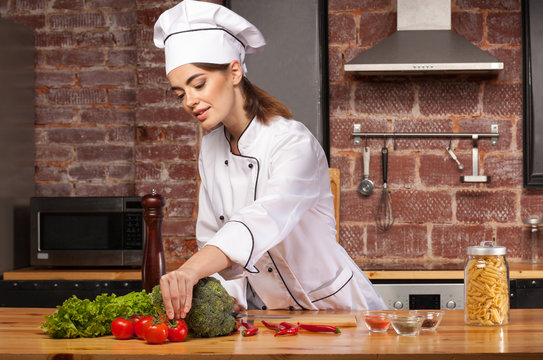Female cook in a white hat in the kitchen preparing a meal with pasta, salad, broccoli, tomatoes and pepper