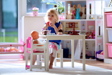 Cute preschooler girl having birthday tea party with her doll. Little child plays in sunny room at home or kindergarten. Kids using toy dishes and cakes.