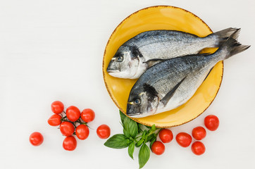 Raw fresh dorado fish on yellow plate, cherry tomatoes, basil. White table, top view, copy space. 