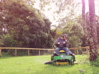 A baby boomer is mowing grass on a rural property on a ride on lawn mower. Filtered image for a...