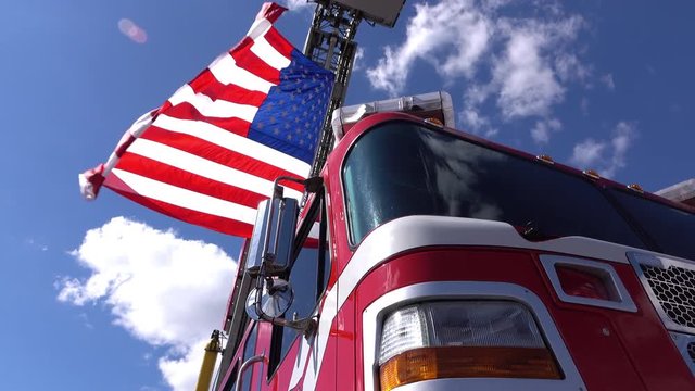 Firetruck sits beneath blue sky with ladder extended as American flag waves in the wind.

