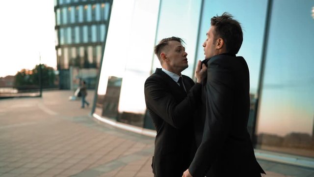 Angry Young businessman grabbing his colleague by the lapels of suite. He want to start fighting. Hard conversation and debate. Boss look at smart phone. Glass business centre building sunset at the