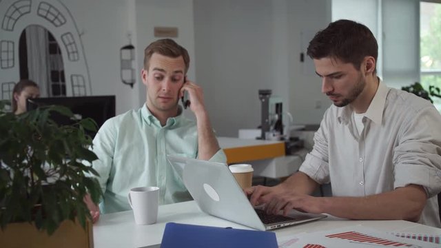 Teamwork professional men in the startup company. Caucasian handsome man talking on the smartphone. Mixed race male typing on the computer. On the table financial documents with diagrams charts.