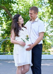 pregnant woman with husband posing in the city park, family portrait, summer season, green grass and trees