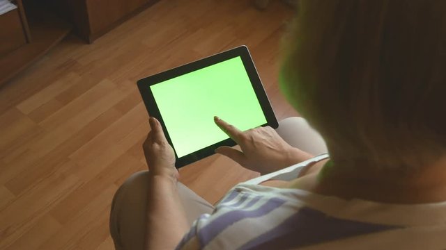 Elderly woman sitting on sofa at home and using a digital tablet pc with green screen, back view. Tablet PC in a adult woman hands 