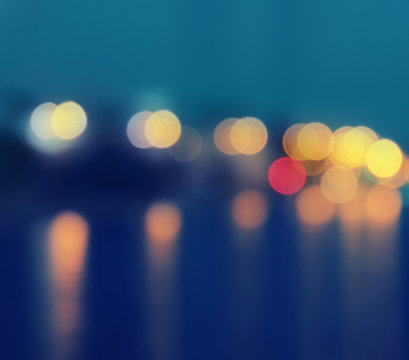 Square image of a blurred city lights with bokeh effect reflected on water.