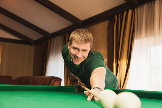 Young man playing billiards. Cheerful guy smiling at the camera. It keeps in the hands of the cue. Green billiards table.