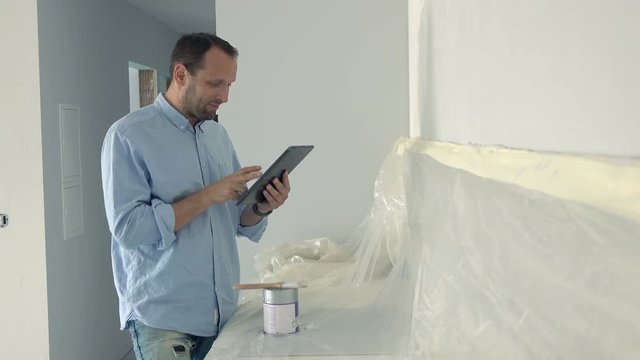 Young man standing with tablet during painting wall in his new home, 4K
