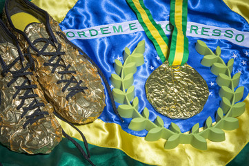Golden running shoes resting with gold medal and laurel wreath on shiny Brazilian flag still life 