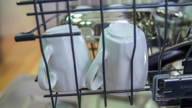 Pushing the top shelf with white cups inside a dishwasher and then slowly closing the dishwasher before turning it on. Close-up shot
