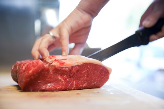 Close Up Of Butchers Hands Slicing Raw Steak On Butchers Block