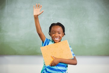 Happy schoolboy raising his hand and holding books in class room