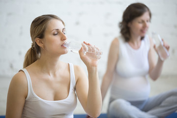 Pregnancy Yoga, Fitness concept. Portrait of two beautiful young pregnant yoga models drinking water with closed eyes after working out indoor. Group of pregnant fitness women resting in fitness class