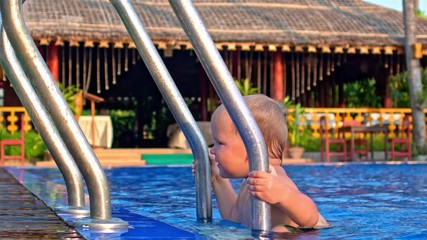 toddler boy climbing out of a outdoor swimming pool with ladder on tropical resort background