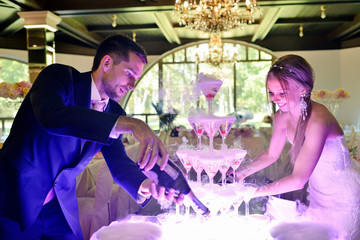 Beautiful wedding couple is pouring champagne indoors. Colorful glasses for alcohol with berries for bride and groom. Beauty of bridal interior for marriage. Bright bar for celebration