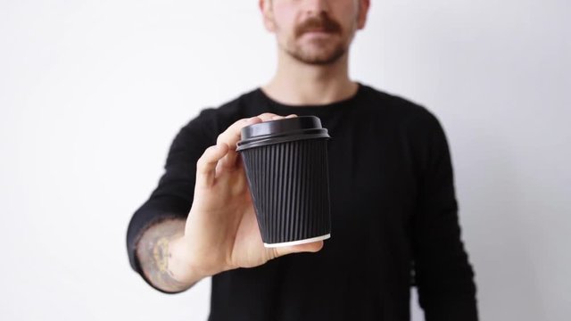 Handsome bearded in unfocused bokeh guy shows focused black unlabeled paper cup on camera