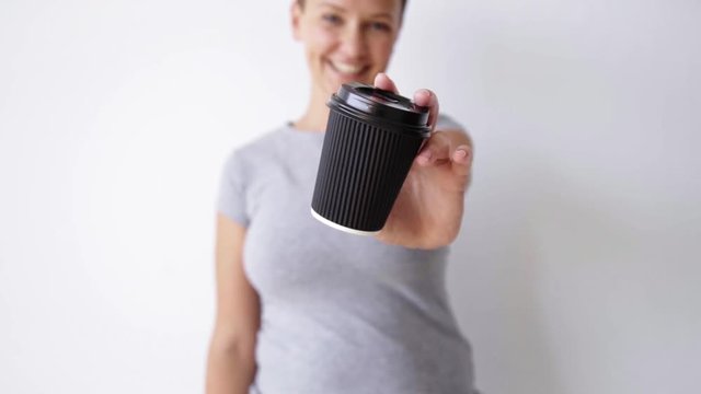 Unfocused handsome girl in blank shirt shows unlabeled black paper cup on camera smiling