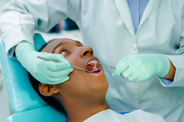 Young hispanic man lying in chair receiving dental treatment with mouth open, dentist hands wearing...
