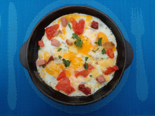 Freshly made scrambled eggs with sausage and tomatoes in a pan, which stands on a wooden stand and blue tablecloths 