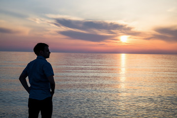 Young man standing at the beach in front of amazing sea view at sunset or sunrise and thinking about his future. Rear view