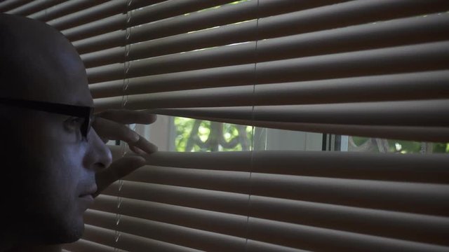 30s  Man in the glasses looks and searches  looks out  through the blinds in the window. The concept of fear, paranoia, phobia, fetish.In a dark room.