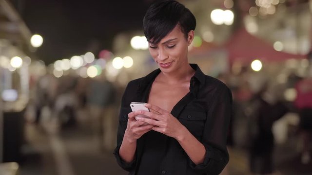 Beautiful millennial woman using smartphone texting app and making a phone call at outdoor evening farmers market. Happy smiling black woman in her 20s.