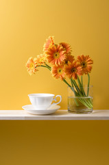 黄色の背景とオレンジ色のガーベラと花瓶とコーヒーカップ