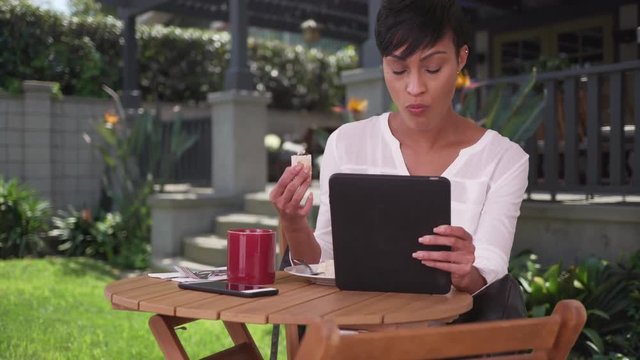 Black woman sitting at table in her backyard eating lunch and using tablet computer. African American millennial homeowner drinking coffee with lunch.