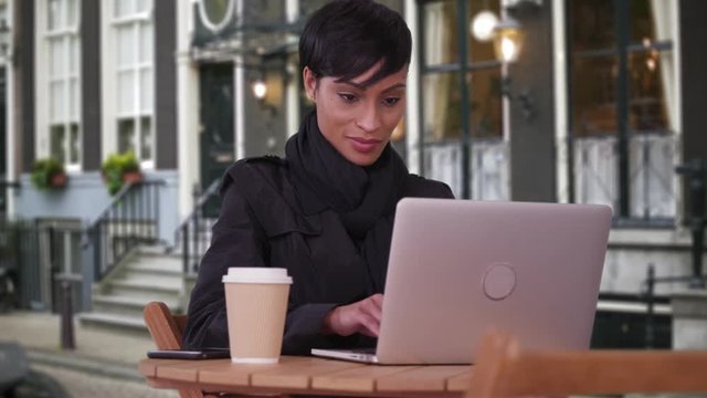 Fashionable woman using laptop computer at outdoor café in Amsterdam. Happy smiling millennial in black overcoat on urban European street typing.