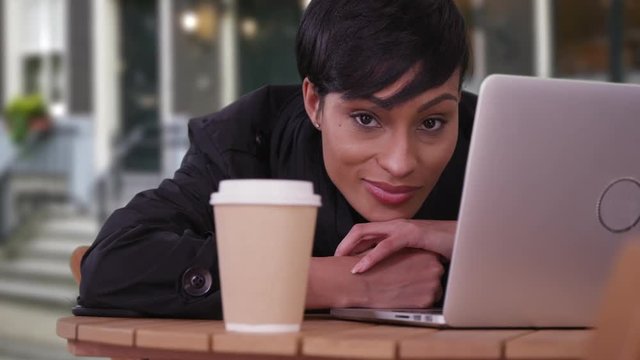 Happy smiling professional black woman at outdoor café in Amsterdam. Close up of woman with laptop computer and coffee cup. Beautiful millennial woman in her 20s on urban European street.
