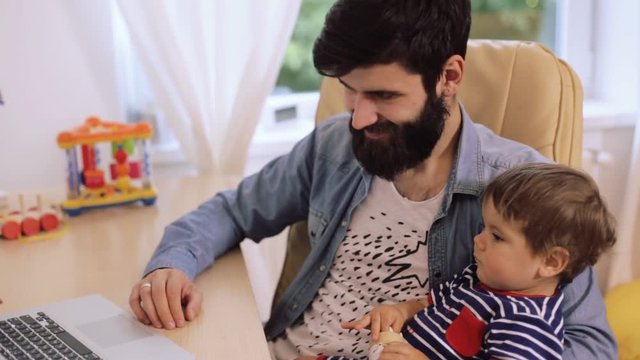 Family time. Young smiling bearded father and his cute son using computer together at home