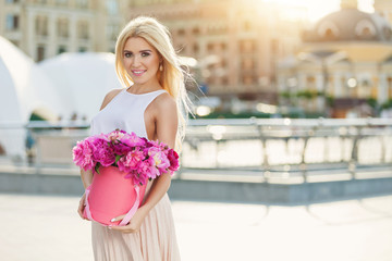 Obraz na płótnie Canvas Portrait of young beautiful blonde woman with box of flowers posing in the city streets. flower box and gift box. bouquet of flowers in gift box. birthday, March 8, Valentine's Day, romantic