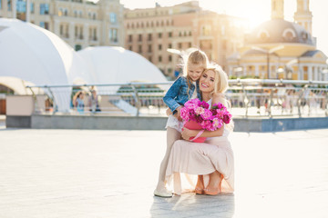 Obraz premium Portrait of young beautiful mother and daughter with box of peonies posing in the city streets. Flower box and gift box. Bouquet of flowers in gift box. Birthday. March 8