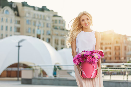 Portrait of young beautiful blonde woman with box of flowers posing in the city streets. flower box and gift box. bouquet of flowers in gift box. birthday, March 8, Valentine's Day, romantic