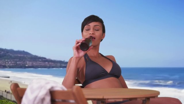 Woman on vacation drinking bottle of beer and enjoying the beach sun. Happy smiling black woman in her 20s sitting at resort table.