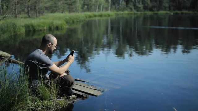A man sits on a pier of the lake and taking pictures on the phone. The beautiful blue lake and forest on background