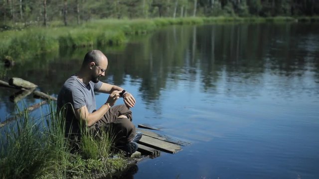 A man sits on a pier of the lake and touch smartwatch. The beautiful blue lake and forest on background