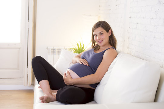 young beautiful happy 8 or 9 months pregnant woman at home living room couch holding big belly