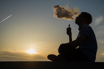 vaping young man with, produces vapor on sunset sky background at the sea coast promenade, place...