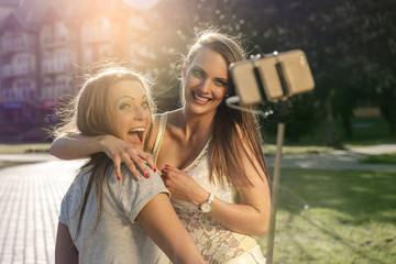 Summer outdoor portrait of two friends fun girls taking photos with a smartphone at bright sunset. Group of happy women taking self-portrait on their travel vacations. Wearing style dress.