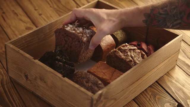 Tattooed chief hand takes one bread from many samples inside the wooden box on aged table and shows on camera to present Professional artisan bakery business with samples.