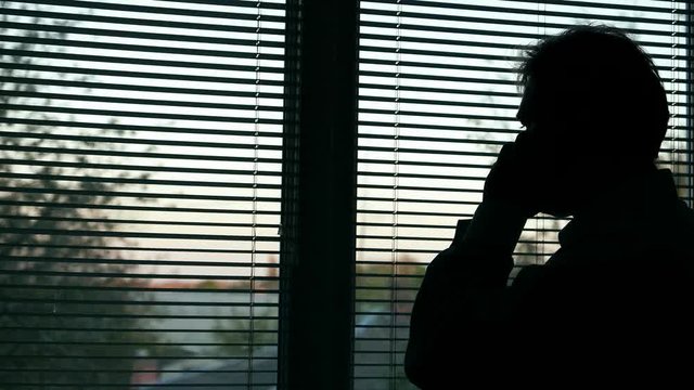 Male man looking out the window and makes an urgent call on the phone