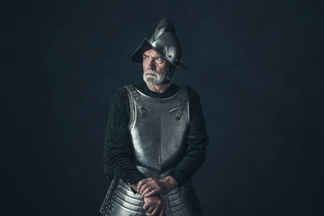 Bearded knight standing with sword in front of gray wall.