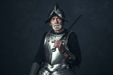 Knight in armor with gray beard holding sword.