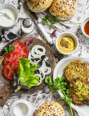 Ingredients for cooking vegetarian burgers - zucchini burgers, homemade buns, tomatoes, onions, herbs, spices and sauces on a light stone background. The view from the top