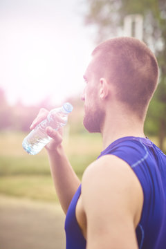 Tired man drinking water from a plastic bottle after fitness time and exercising in city street park at beautiful summer day. Sporty model training outdoor.