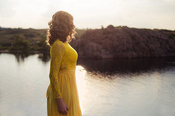 Young woman staying back. Attractive girl enjoys sunset near river and cliff in summer light