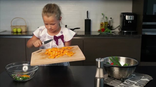 Little girl pouring yellow bell pepper into a bowl for the salad in the kitchen